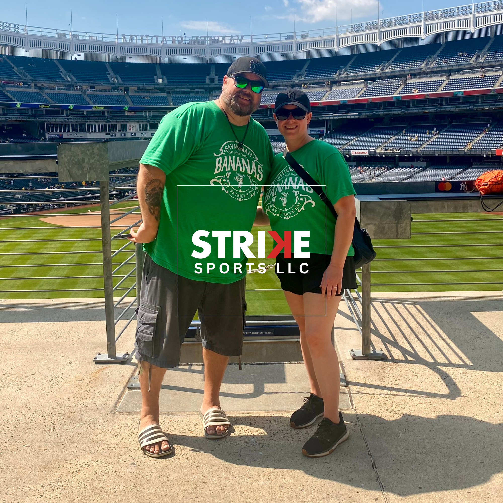 A couple in matching green shirts stands in front of the field at Yankee Stadium from the promenade.